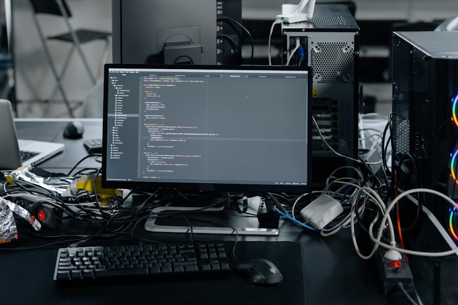 The image depicts a cluttered workspace featuring a computer monitor displaying lines of code, surrounded by tangled cables, a keyboard, and a computer mouse. The background includes computer towers w