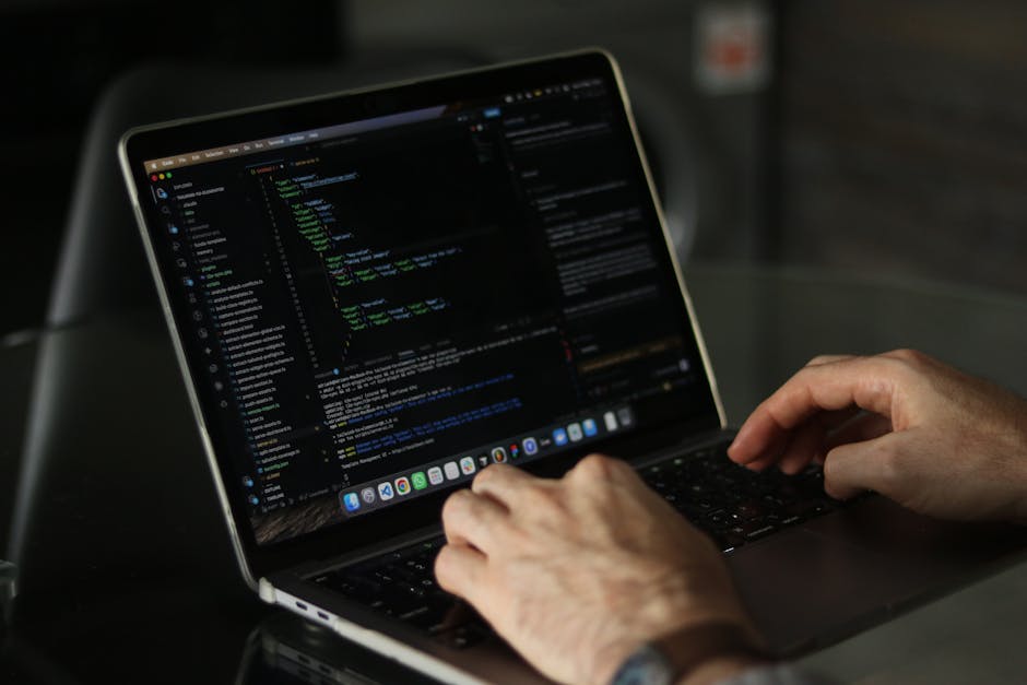 A close-up of a person's hands operating a laptop computer on a dark, reflective surface. The laptop screen displays a code editor with colorful syntax highlighting in a dark theme, and various lines 